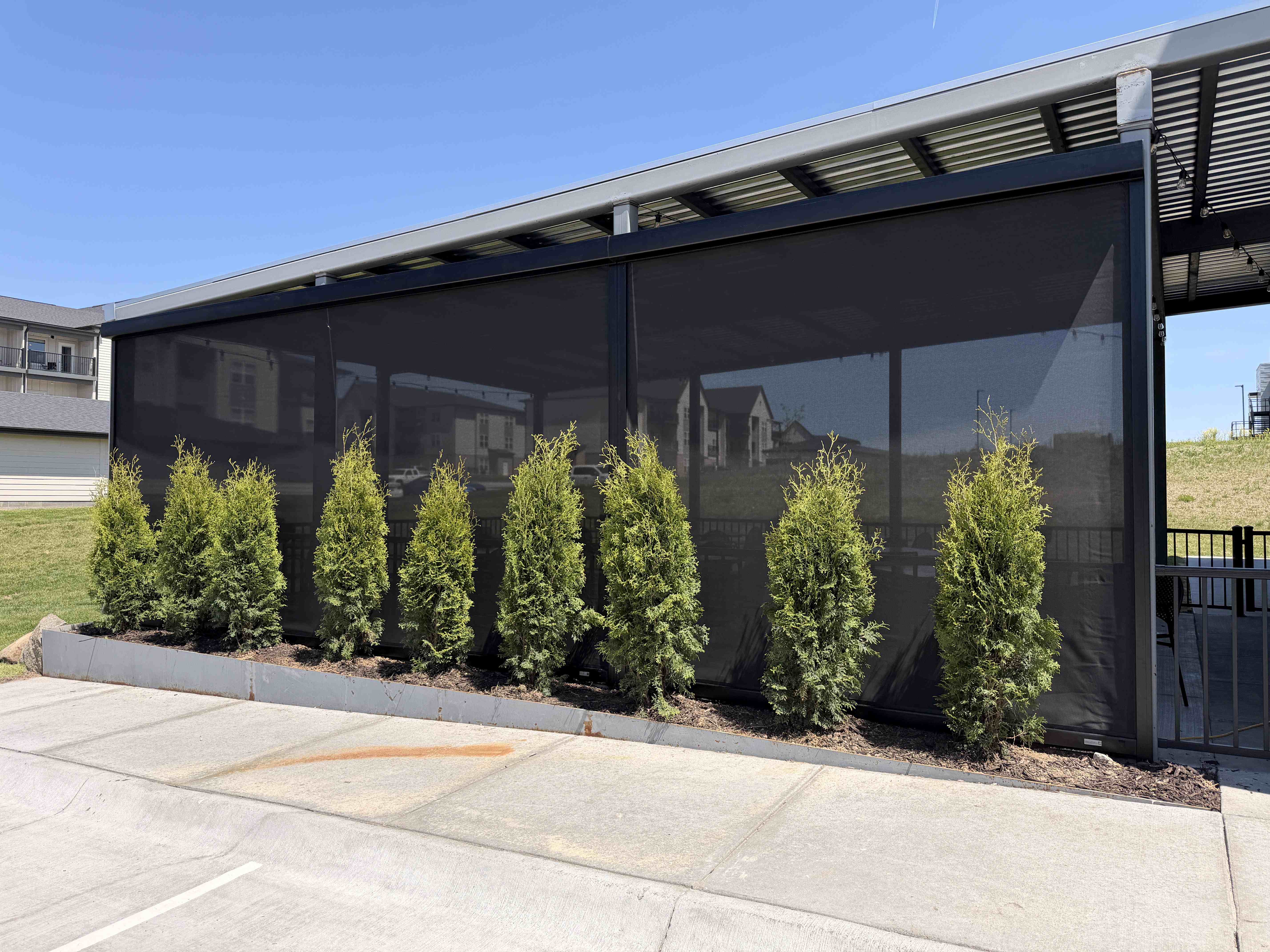 Commercial pavilion enclosed with dark motorized screens behind a row of evergreen shrubs