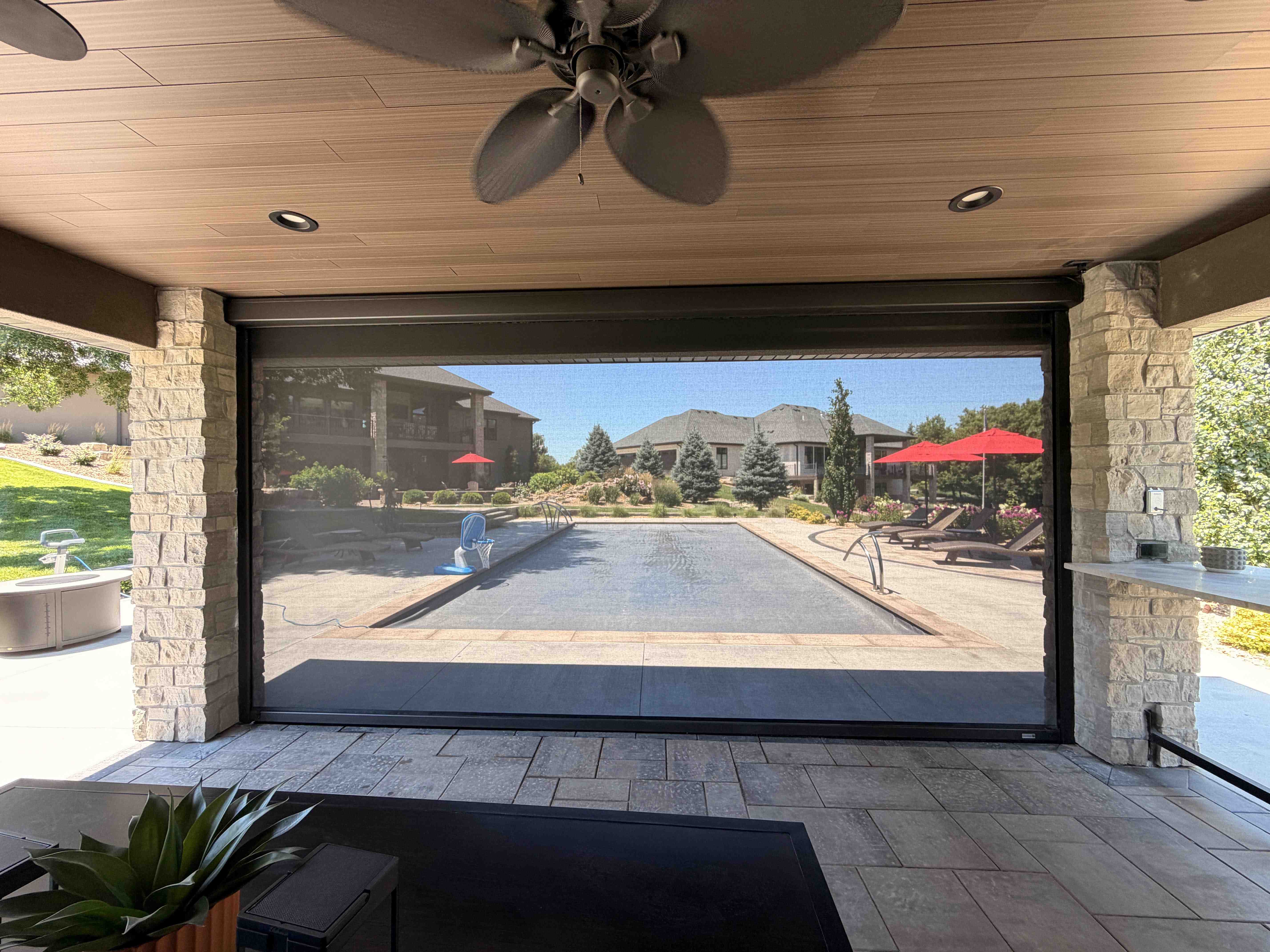 View of a backyard pool through a lowered motorized screen on a covered stone patio